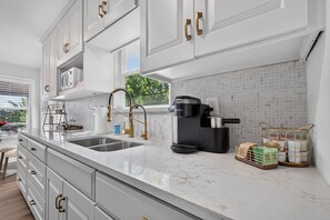 A modern kitchen with white cabinetry, a double sink, a coffee maker, and a window with a view of greenery. - New! Cozy home, Game Room + Covered Porch (Austin)