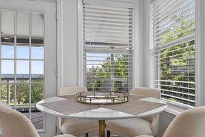 Ein Zimmer mit einem runden Tisch, vier Stühlen und Blick auf Grün durch große Fenster mit Jalousien.