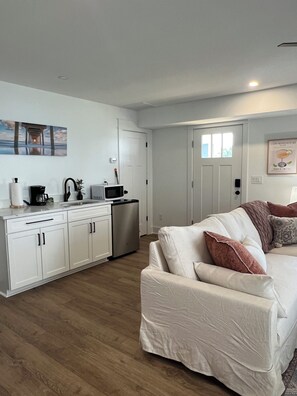A modern kitchen with white cabinets, a stainless steel refrigerator, and a microwave. There is a white sofa with brown cushions in the living area. - Water Views and Great Location - BRAND NEW - 3 bedroom 3 bath soundside cottage (Kill Devil Hills)
