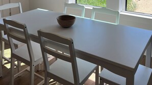 A dining area with a white table and chairs, a hanging light fixture, a framed picture, and large windows with a view of greenery and a building. - Water Views and Great Location - BRAND NEW - 3 bedroom 3 bath soundside cottage (Kill Devil Hills)