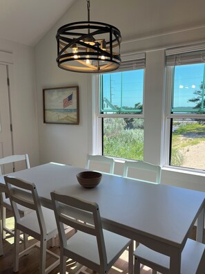 A dining area with a white table and chairs, a hanging light fixture, a framed picture, and large windows with a view of greenery and a building. - Water Views and Great Location - BRAND NEW - 3 bedroom 3 bath soundside cottage (Kill Devil Hills)