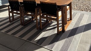 An outdoor wooden table and chairs under a covered area with a view of a beach and a house in the background. - Water Views and Great Location - BRAND NEW - 3 bedroom 3 bath soundside cottage (Kill Devil Hills)