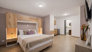 Un dormitorio moderno con una cama, mesitas de noche, un televisor y un baño visible al fondo.
