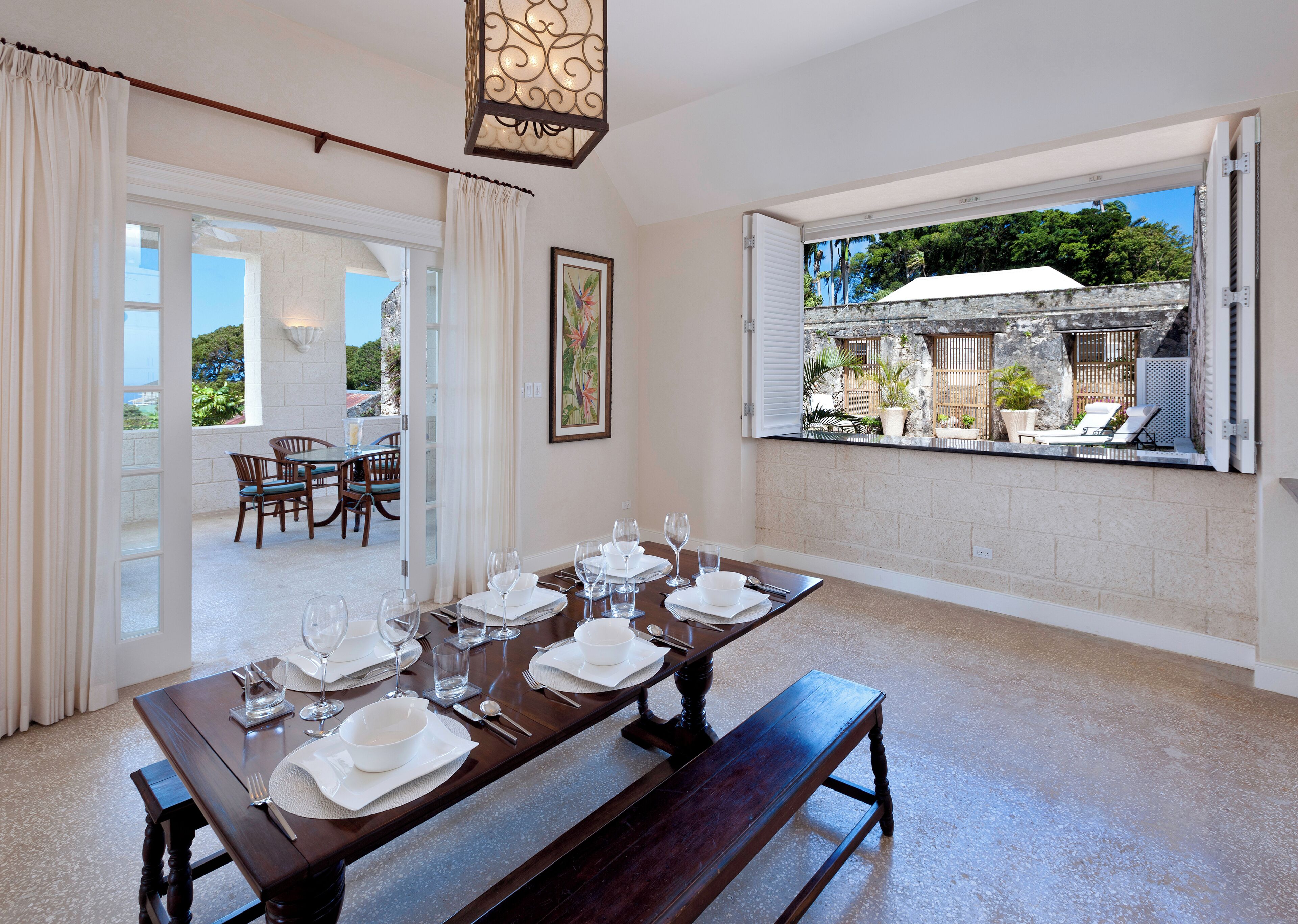 Un coin repas avec une table dressée pour six personnes, un banc et une vue sur un patio extérieur agrémenté d’un parasol blanc.