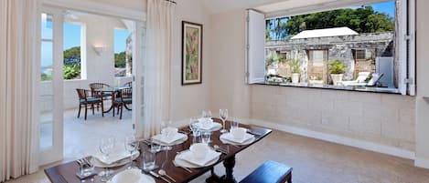 Un coin repas avec une table dressée pour six personnes, un banc et une vue sur un patio extérieur agrémenté d’un parasol blanc.