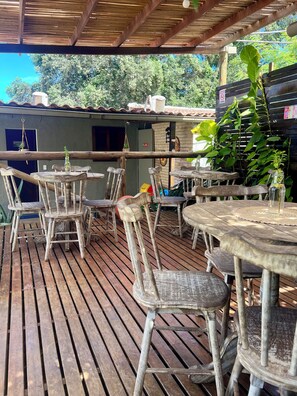 An outdoor patio with wooden tables and chairs, a covered area, and a brick building in the background.