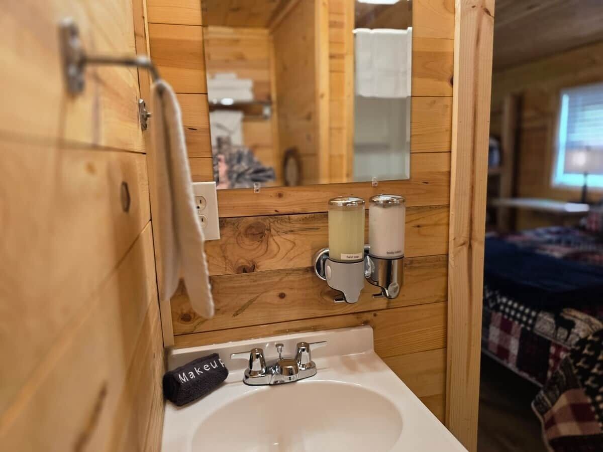 A wooden cabin-style bathroom with a sink, mirror, towel rack, and two soap dispensers.