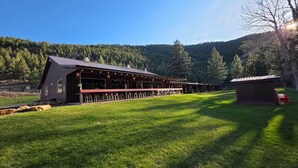 Ein großes, stallartiges Gebäude mit einer überdachten Außenlounge, umgeben von einer grünen Wiese und bewaldeten Hügeln.