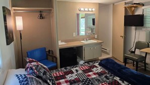 Quarto compacto com cama, escrivaninha com espelho, cadeira e televisão.