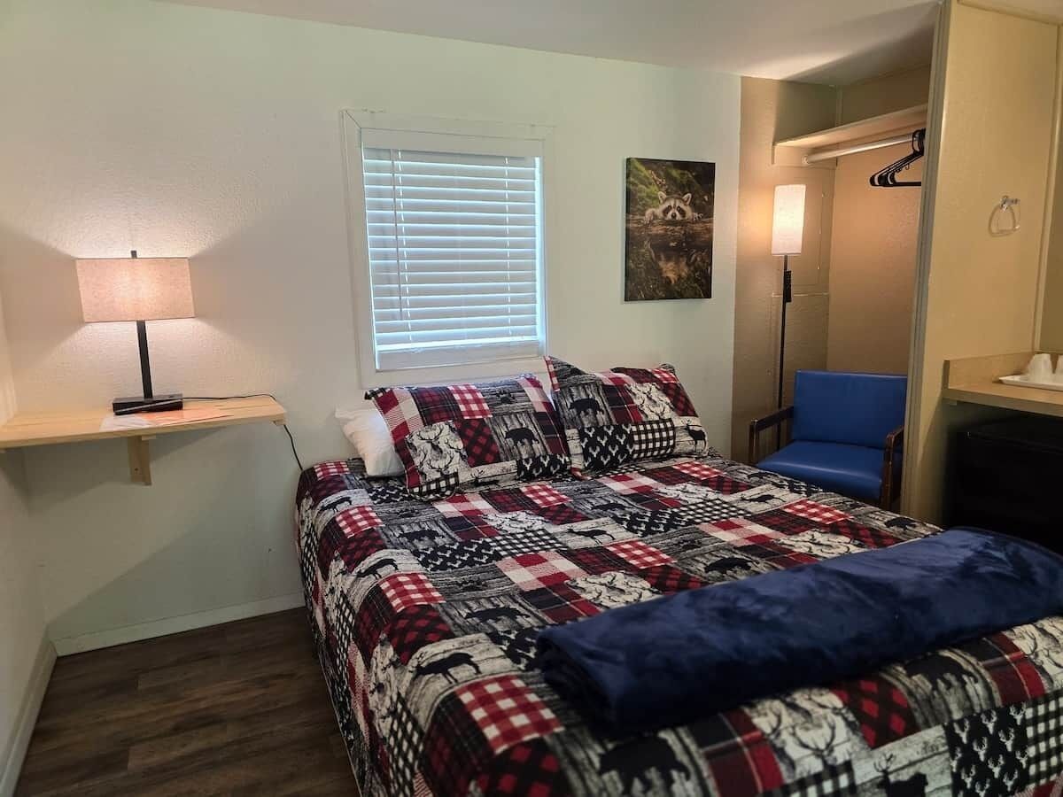 A bedroom with a bed, a nightstand, a lamp, a window with blinds, a chair, and a door leading to another room.