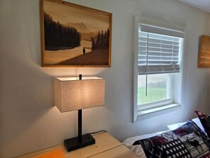 Um quarto com uma cama, uma mesa de cabeceira com abajur, uma janela com persianas e um quadro na parede.