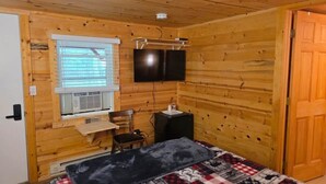 Ett rum med träpanel på väggarna, innehållande en säng, ett skrivbord, en stol, en tv och ett fönster med persienner.