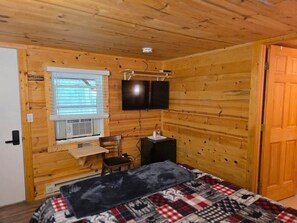 Une pièce aux murs lambrissés, comprenant un lit, un bureau, une chaise, un téléviseur et une fenêtre avec des stores.