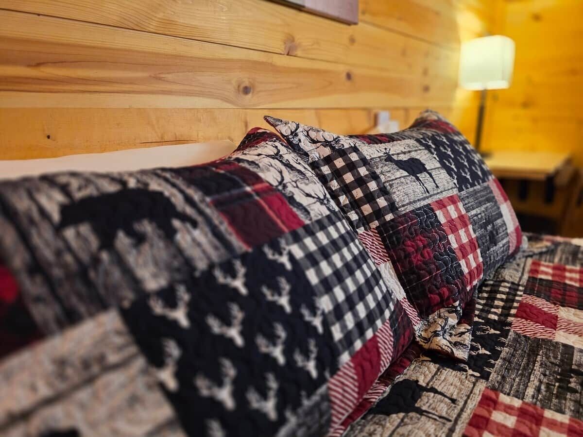A bed with a patterned quilt and pillows, wooden headboard, and a lamp in the background.