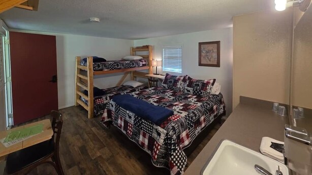 Una habitación con literas, un escritorio, una silla, una cama con manta estampada, una ventana con persianas y un baño con lavamanos y espejo.