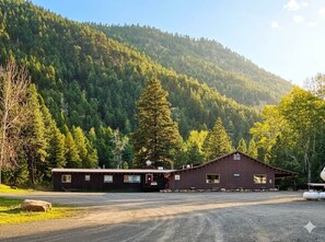 Un edificio rustico con vialetto sterrato, immerso in una fitta foresta e con montagne sullo sfondo.