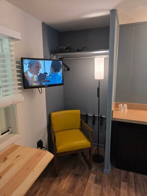 Habitació petita amb televisió penjada a la paret, una cadira groga, una taula de fusta i una petita zona de cuina.