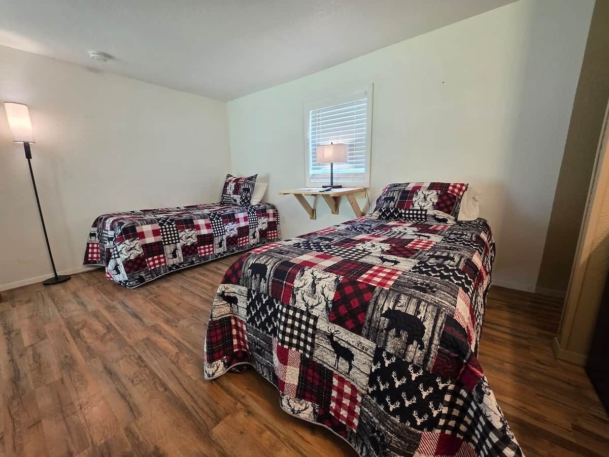 A room with two beds, a standing lamp, a window with blinds, and a small table.