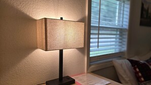 Una lámpara de mesa sobre una mesita de noche de madera, junto a una ventana con persianas.