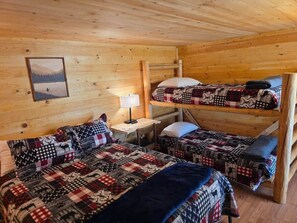A cabin-style room with bunk beds, a wooden ceiling, a framed picture on the wall, and a small table with a lamp.