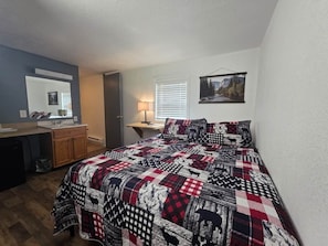 Ein Schlafzimmer mit einem Bett, einem Schreibtisch, einem Stuhl, einem Spiegel und einem Fenster mit Jalousien.