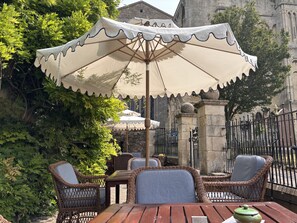 Un espace extérieur aménagé avec des chaises et une table en osier, un grand parasol, et un bâtiment en pierre en arrière-plan.