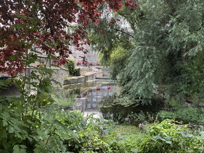 A serene riverside view with a stone bridge, lush greenery, and a calm water body reflecting the surroundings.
