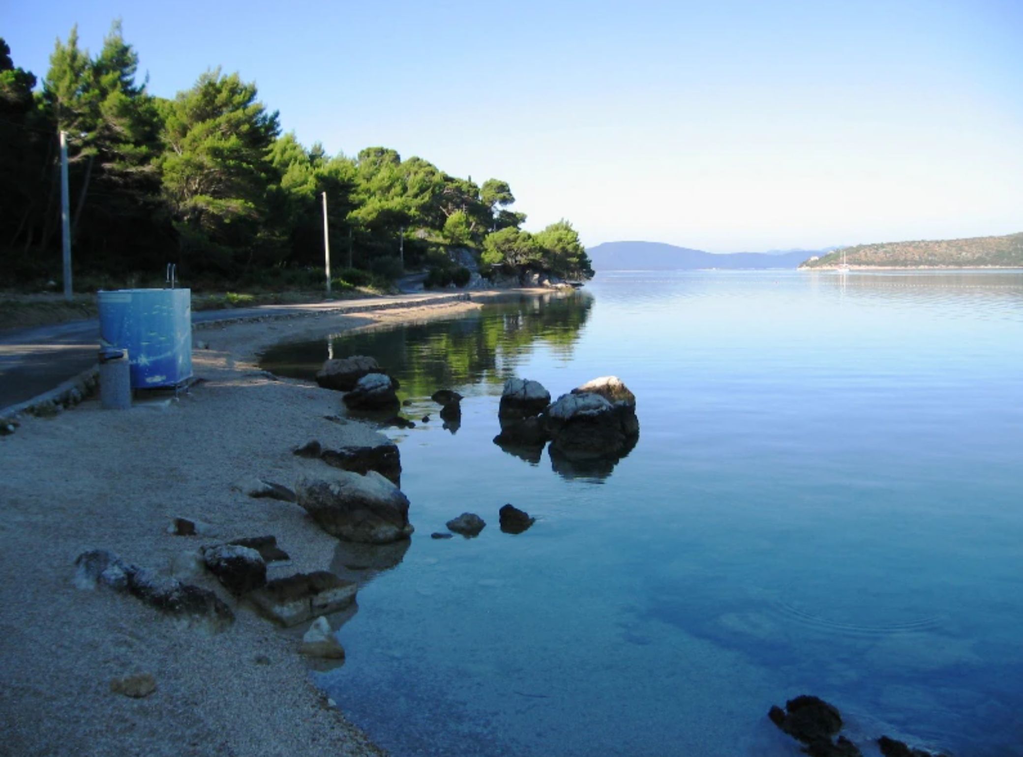 A calm lake with a rocky shoreline, trees, and a clear sky.