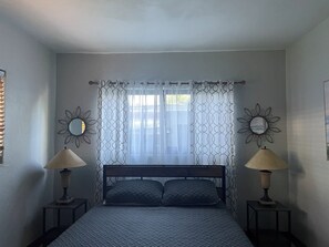 Ein Schlafzimmer mit einem Bett, zwei Nachttischlampen, einem Fenster mit gemusterten Vorhängen und dekorativen Wandspiegeln.