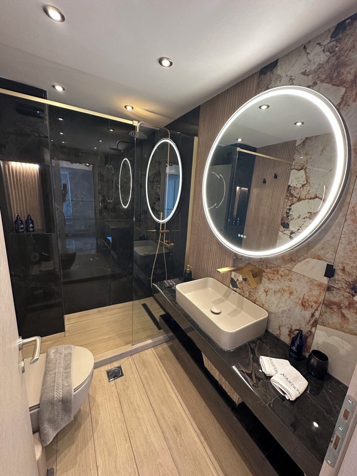 A modern bathroom with a glass shower enclosure, a large vanity with a round mirror, and a marble wall.