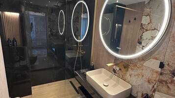 A modern bathroom with a glass shower enclosure, a large vanity with a round mirror, and a marble wall.