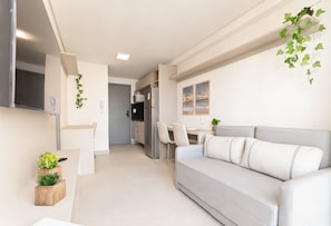 Ein modernes Wohnzimmer mit einer Couch, einem Esstisch und Stühlen. Es gibt eine Küchenzeile mit einem Kühlschrank und einer Mikrowelle.