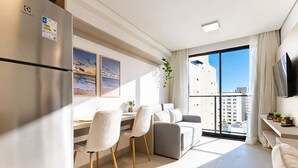 A modern living room with a dining area, a large window with a view, a flat-screen TV, and a refrigerator. - AT Self #804 Steps From the Beach by Carpediem (João Pessoa)