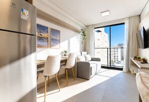 A modern living room with a dining area, a large window with a view, a flat-screen TV, and a refrigerator. - AT Self #804 Steps From the Beach by Carpediem (João Pessoa)