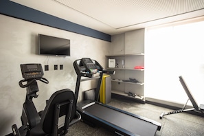 Ein Fitnessraum mit Laufband, Crosstrainer und Ergometer, einem Fernseher an der Wand und einem Fenster mit Tageslicht.