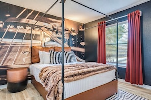 Ein Himmelbett mit Felldecke, ein runder Holzschemel und ein Wandgemälde.