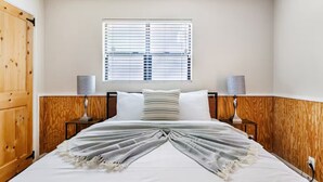 A bedroom with a wooden wardrobe, a bed with a white and gray blanket, two bedside lamps, and a window with blinds. - 3-bedroom Phoenix Cabin with cool AC for relaxing stay (Phoenix)