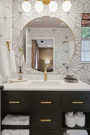 Ein Badezimmer mit einer Wand mit geometrischem Muster, einem runden Spiegel, einem goldenen Wasserhahn und einem dunklen Holzwaschtisch mit Schubladen.