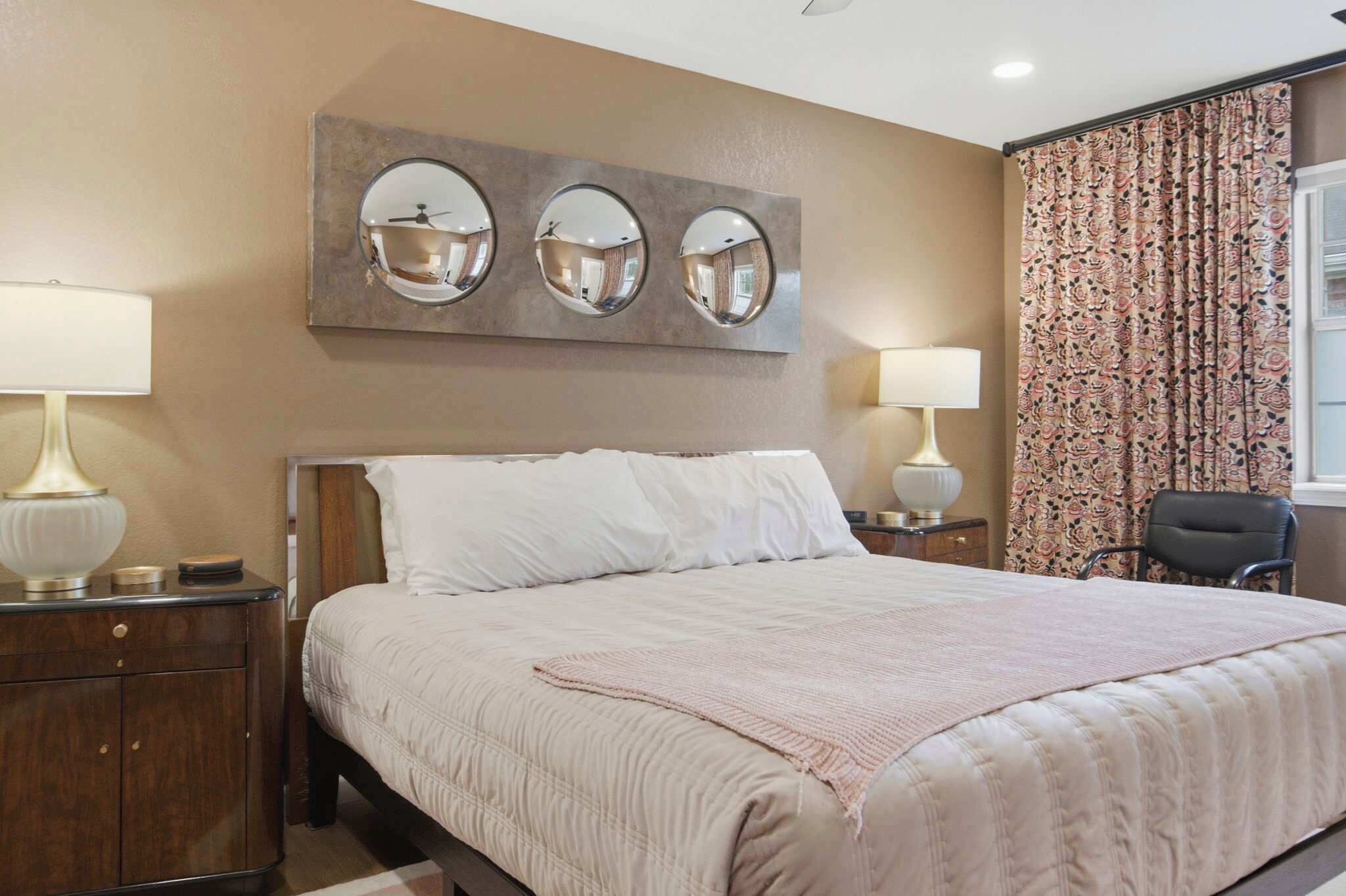 Habitación de hotel con cama, dos mesitas de noche con lámparas, un espejo, una silla y una ventana con cortinas.