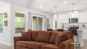 A modern living room with a brown sofa, a dining area with a bar, and a kitchen with white cabinets and a microwave. - May Way | Trails, Golf, Hiking & Relaxation (Bella Vista)