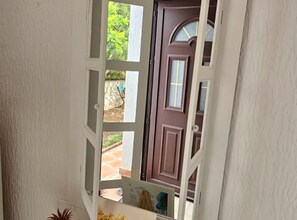Blick durch eine Tür zu einer braunen Tür mit Fenster, einer weißen Wand und einem kleinen Außenbereich mit einer Pflanze.