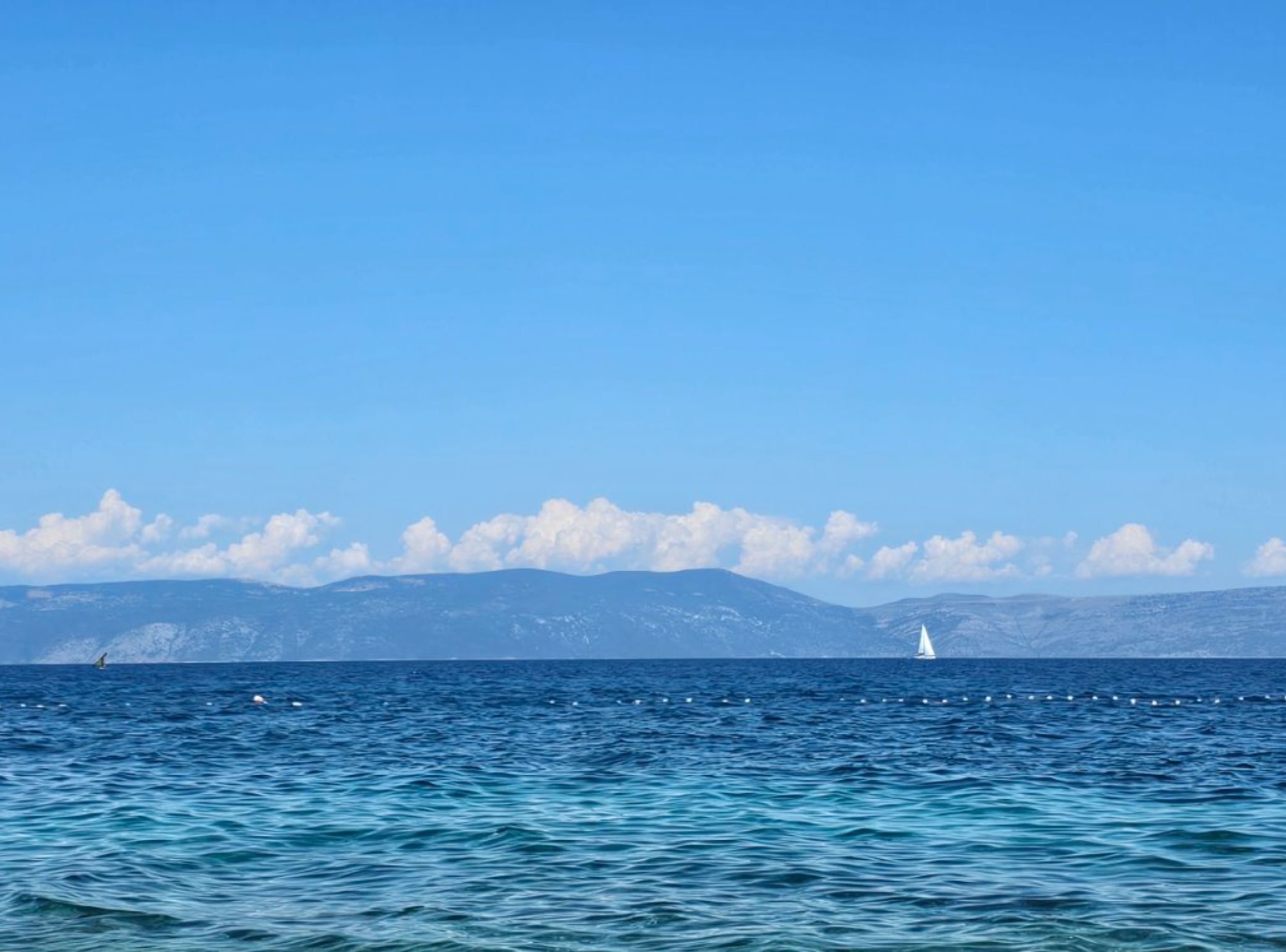 A clear view of the sea with a sailboat in the distance and mountains in the background.