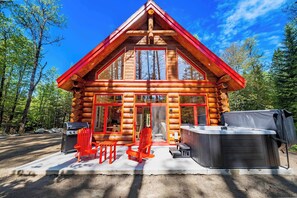 A log cabin with a large front door, two red chairs, a hot tub, and a grill.