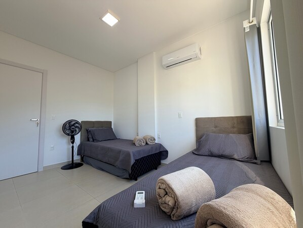 A bedroom with two beds, a standing fan, an air conditioning unit, and a window.