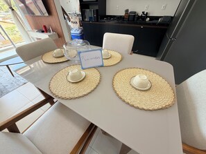 A dining table with four placemats and teacups, a sign reading 'Bem-vindo!', and a kitchen in the background.
