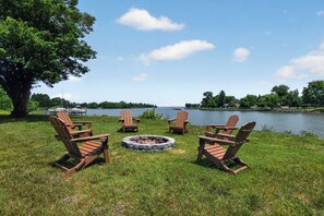 Eine Uferlandschaft mit vier Adirondack-Stühlen, die um eine Feuerstelle aus Stein angeordnet sind, einem großen Baum und Blick auf einen See mit Booten und Häusern im Hintergrund.