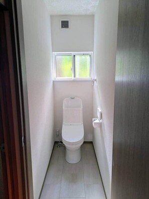 A narrow, white-walled bathroom with a toilet, a window above it, and a door on the right. - Yuki Niwa (Myoko)