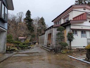 A traditional Japanese building with a wooden exterior, a stone pathway, and a small garden with various plants and a stone lantern. - Yuki Niwa (Myoko)