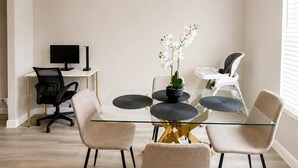 Ett modernt matområde med ett glasbord, fyra stolar, en liten krukväxt och ett skrivbord med en dator.