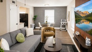 Ein Wohnzimmer mit grauem Sofa, einem braunen Sessel, einem kleinen runden Tisch, einem Fernseher und einem großen Fenster mit Blick auf die Berge.